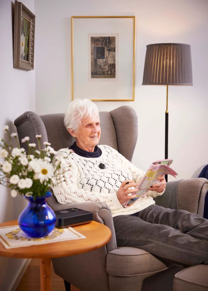 Oudere vrouw zit thuis op een stoel met alarmketting om en personenalarmering basisstation op de tafel
